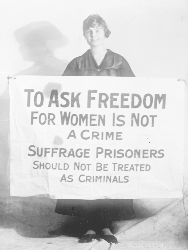Schwarz-weißes Foto einer Frau, die ein Transparent hält, auf dem steht: "Für die Freiheit der Frauen zu kämpfen ist kein Verbrechen, Suffragetten sollten nicht wie Kriminelle behandelt werden" und dabei lächelt.