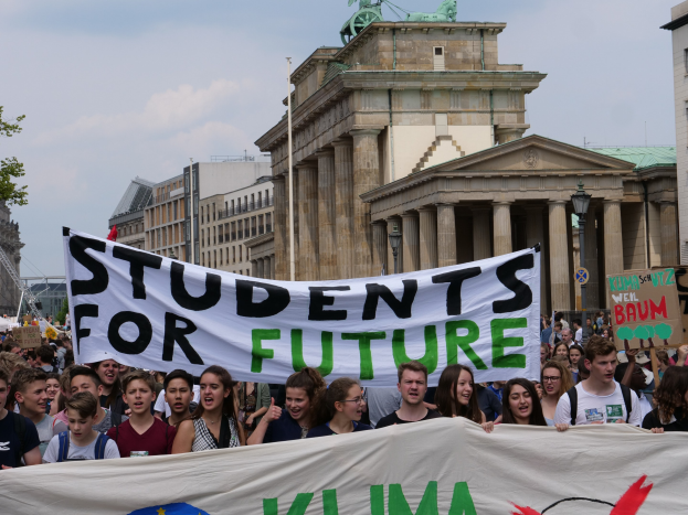 Gruppe von Schülern marschiert in Berlin mit einer leuchtend bunten "Students for Future"-Schlagzeile an Gebäuden, Bäumen und Himmel vorbei.