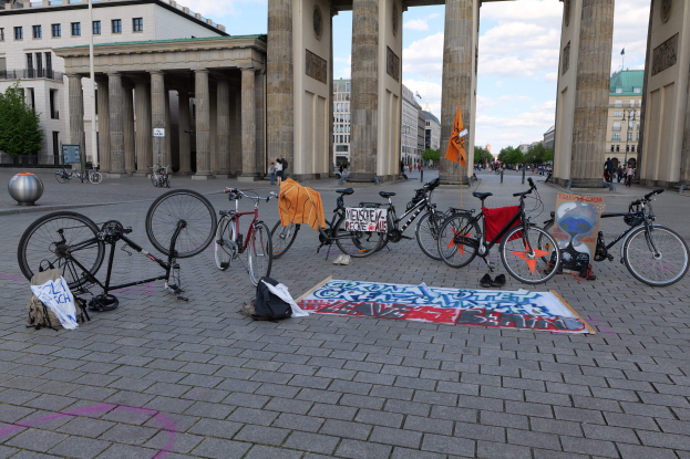 Eine Gruppe von Fahrr├Ądern, die vor dem Brandenburgertor in Berlin, Deutschland, geparkt sind, mit Taschen und einer Fahne in der N├Ąhe, vor einer Kulisse aus Geb├Ąuden, B├Ąumen und einem klaren blauen Himmel.