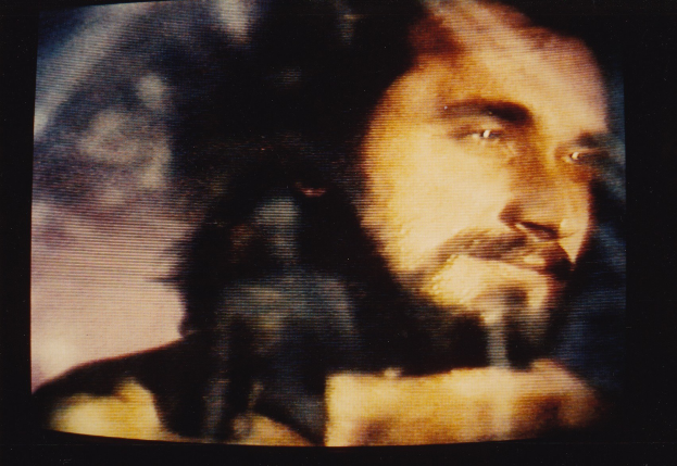 Ein Mann mit langem dunklen Bart und Haar, der ein weißes Hemd trägt, erscheint auf einem Fernseher mit dunklem Hintergrund und schaut direkt in die Kamera mit einem ernsten Gesichtsausdruck.