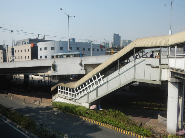 Ein Fußgängerbrücke überspannt eine Straße mit Menschen, die darauf gehen, flankiert von Gebäuden, Straßeninfrastruktur, fahrenden Kraftfahrzeugen und Grün unter einem klaren blauen Himmel.