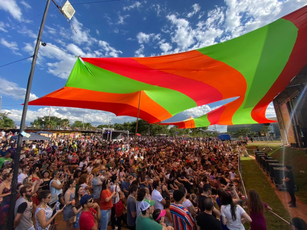 Eine große Menschenmenge steht vor einer Regenbogenflagge mit einem Gebäude, Lautsprechern auf der Bühne und Bäumen im Hintergrund während einer Festival-Jubiläumsfeier.