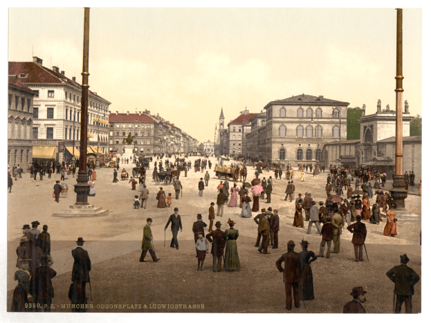 Eine belebte Stadtstraße in München, Deutschland, aus den frühen 1900er Jahren, mit Menschen, die spazieren gehen, einige mit Schirmen, und einem Pferdewagen im Vordergrund, Gebäuden, Bäumen und Polen im Hintergrund und Text unten auf dem Bild.