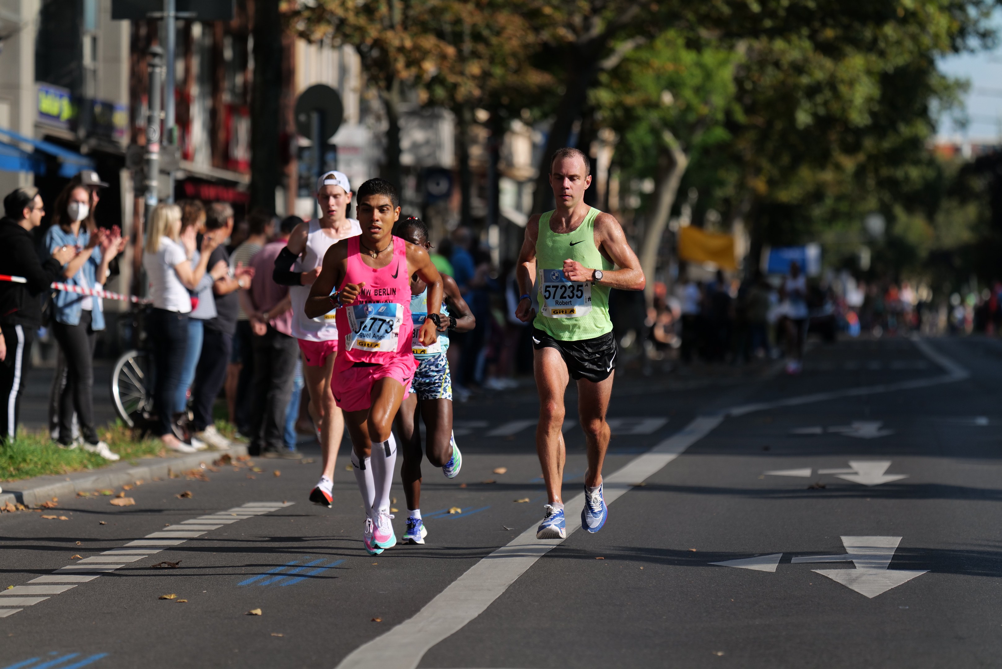 Gruppe von Läufern bei einem Marathon auf einer Stadtstraße mit Zuschauern, Bäumen, Gebäuden und einem Fahrrad im unscharfen Hintergrund.