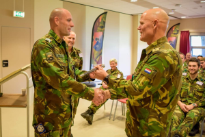 Zwei Männer in Militäruniformen schütteln sich vor einer sitzenden Gruppe die Hände, mit einem Tisch, Fahnen und diversen Hintergrundelementen einschließlich eines Schildes, eines Fensters und Deckenleuchten, was auf eine deutsche Armeeversammlung hindeutet.