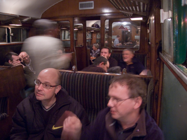 Zwei Männer sitzen auf einem Sitz in einem Zug, mit einer Gruppe von Menschen, die hinter ihnen sitzen; ein Eingang zwischen Abteilen ist oben sichtbar, und ein Fenster ist auf der rechten Seite.