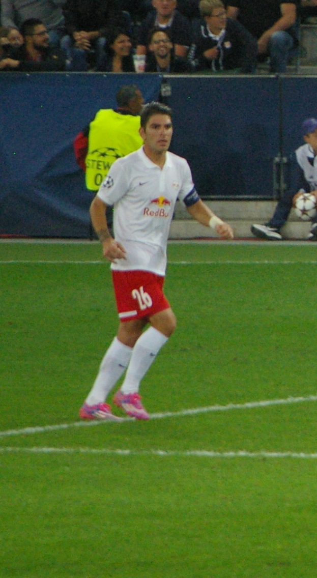 Ein Mann in einem weißen Hemd und roten Shorts, der auf einem Fußballfeld Fußball spielt, mit einer Gruppe von Menschen im Hintergrund.