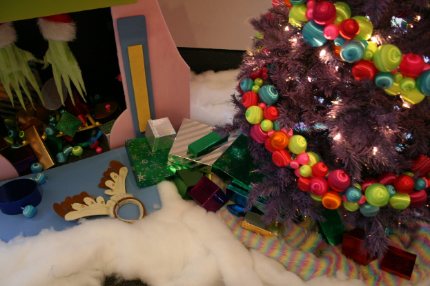 Eine Indoor-Szene mit einer Wand im Hintergrund, einem Leinen- und Wolltuch am Boden, einem geschmückten Weihnachtsbaum rechts, Geschenkboxen und Dekorationsgegenständen in der Mitte und einer Hütte mit Dekorationen oben links.