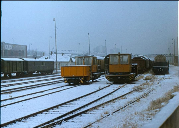 Eine Gruppe von Zügen auf schneebedeckten Schienen mit Pfählen, Gebäuden und einer Brücke im Hintergrund, unter einem sichtbaren Himmel, mit Text am unteren Rand.
