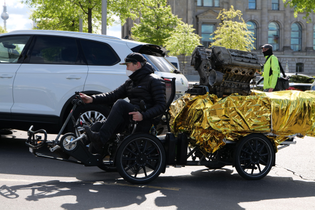 Ein Mann im Rollstuhl mit einem großen Motor auf dem Rücken, umgeben von Fahrzeugen auf einer Straße mit Bäumen, Gebäuden, Polen und einem klaren blauen Himmel im Hintergrund.