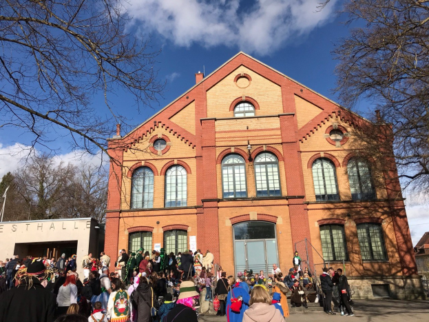 Eine große Gruppe von Menschen, die vor einem mehrstöckigen Gebäude mit Fenstern und Türen steht, umgeben von Bäumen und einem bewölkten Himmel, einige tragen Mützen und tragen Taschen.