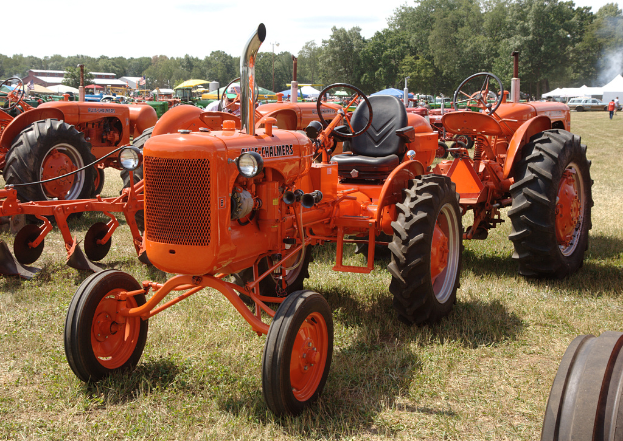 Mehrere orange Traktoren sind auf einem Rasen mit Gras am Boden, Zeltunterstände in Grün, Blau und Weiß, Bäume, Gebäude und Fahrzeuge im Hintergrund und zwei Männer auf der rechten Seite unter einem klaren Himmel.