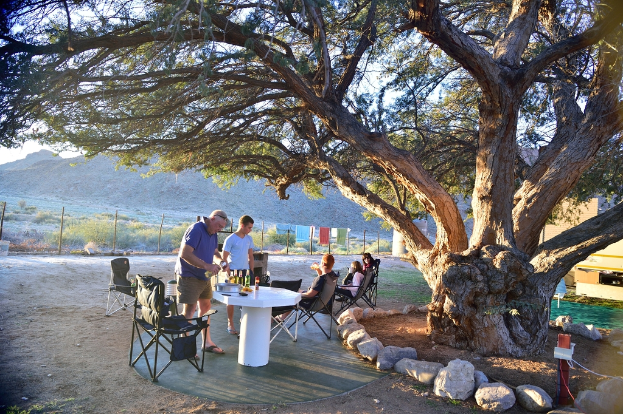 Ein Mann trägt Bierflaschen, während einige Menschen auf Stühlen sitzen, mit einem großen Baum und einem Gebäude im Hintergrund.