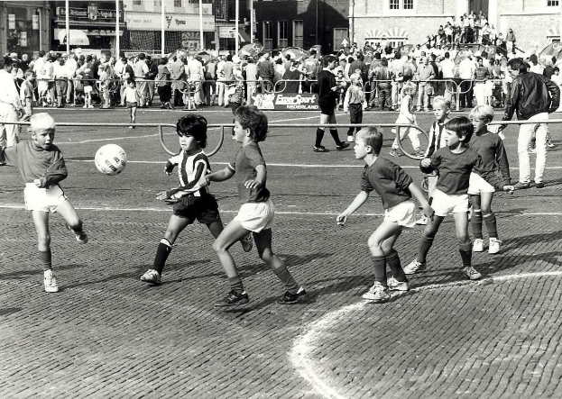 Schwarzes Bild von jungen Jungs, die Fussball auf der Straße spielen, mit einer Zuschauermenge hinter einem Geländer und Gebäuden im Hintergrund.