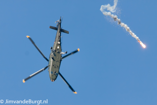 Ein militärischer Hubschrauber im Flug, der eine Rakete hält, die Rauch und Feuer ausstößt, mit Text in der unteren linken Ecke.