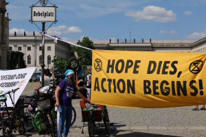 Eine Gruppe von Menschen steht vor einem Gebäude und hält ein gelbes Banner mit der Aufschrift "Hope Dies, Action Begins". In der Nähe stehen mehrere Fahrräder und es gibt einen Pfahl mit einer daran befestigten Tafel. Im Hintergrund sind Bäume und ein klarer blauer Himmel zu sehen.