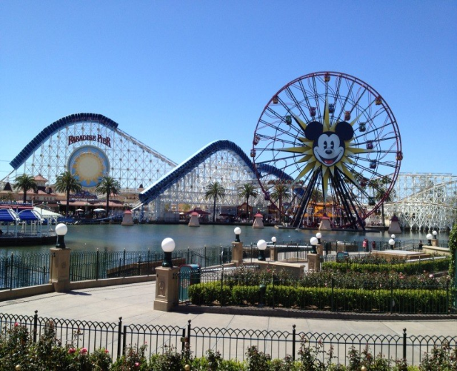 Der Disneyland California Adventure Vergnügungspark mit einem Riesenrad, einer Achterbahn, üppiger Vegetation, Laternenpfählen, einem Wasserbecken im Vordergrund und einem strahlend blauen Himmel.