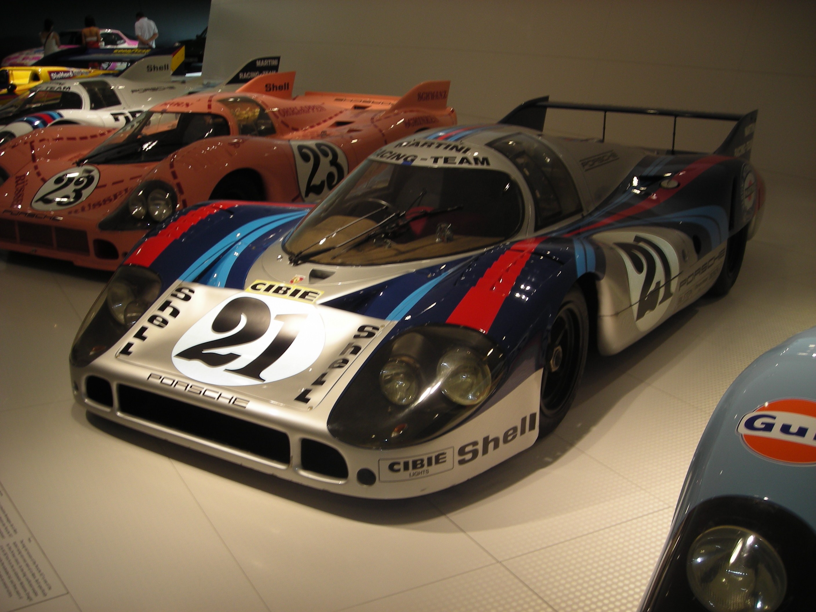 Ein Porsche 917K Rennwagen auf der Le Mans in einem Museum ausgestellt, umgeben von anderen Sportwagen, mit Menschen in der Nähe und einer Wand im Hintergrund; ein Textschild ist links zu sehen.