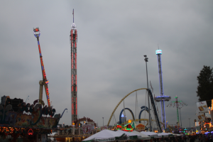 Eine Menschenmenge versammelt sich um einen hell erleuchteten Fasching bei Nacht, mit beleuchteten Fahrgeschäften, Pfählen und Bäumen unter einem bewölkten Himmel.