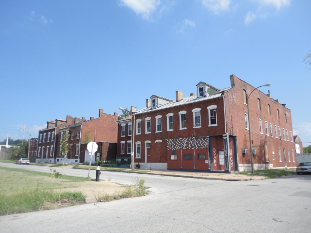 Ein Gebäude mit Pfählen und Straßenlaternen, umgeben von Bäumen und Fahrzeugen auf beiden Seiten, mit einem Gehweg, Gras und einem Feuerlöscher unten und dem Himmel oben.
