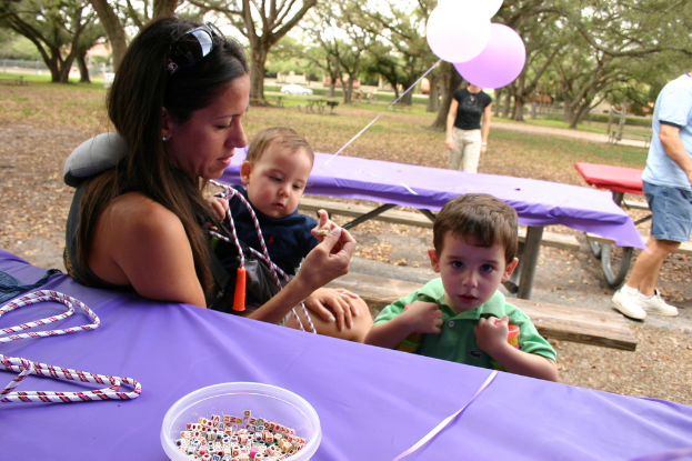 Eine Frau in Schutzbrille hält ein Baby, steht neben einem Jungen in einem grünen Kleid, mit Tischen, einer Violablatt, einer Schale mit Würfeln und Ballons, während ein Mann rechts steht und Bäume im Hintergrund zu sehen sind.