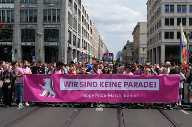 Eine Gruppe von Menschen marschiert auf einer Straße in Berlin und hält ein pinkes Banner mit der Aufschrift "Happy Pride March".