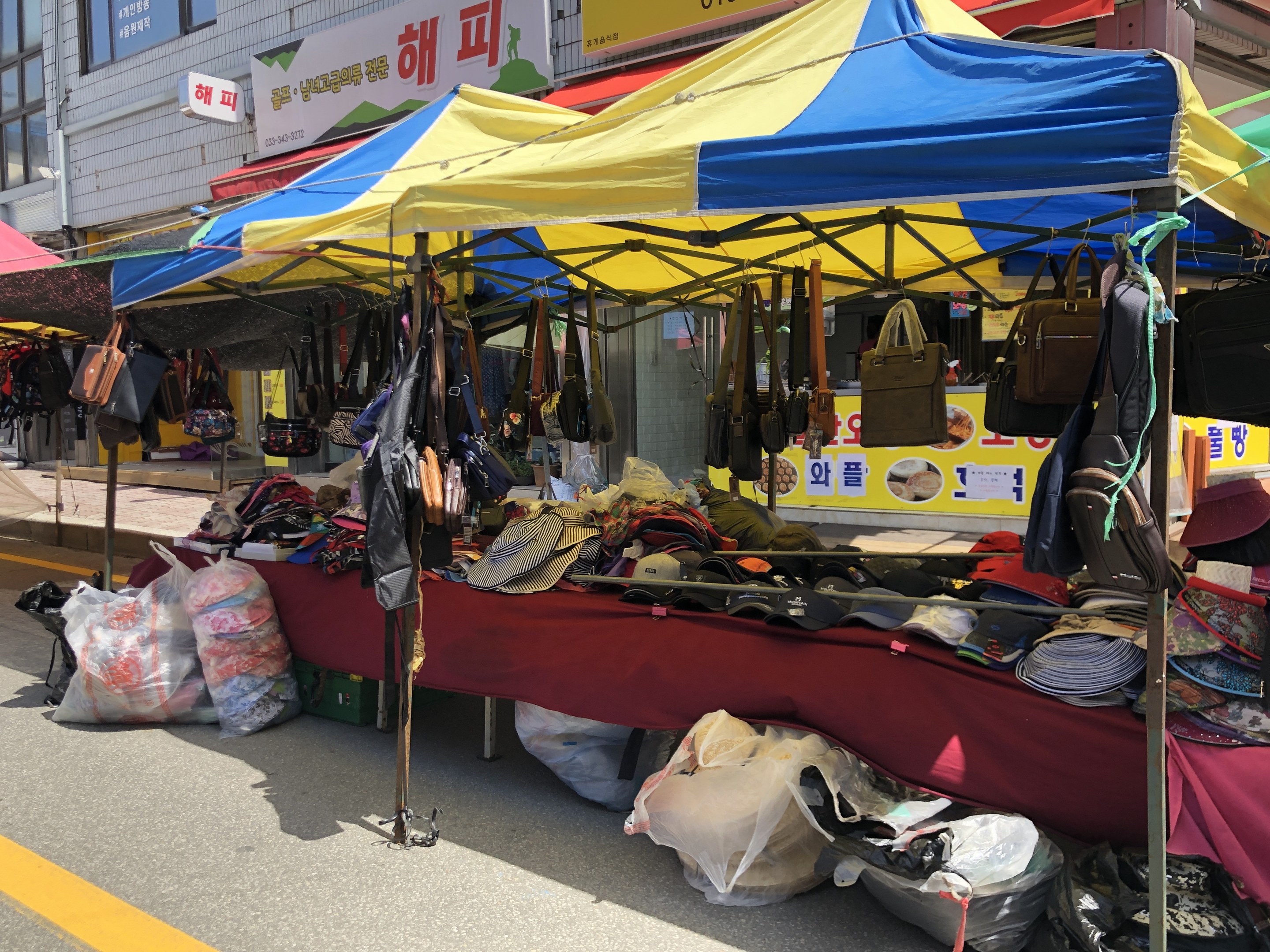 Ein Straßenmarkt mit Zelten, auf denen verschiedene Gegenstände wie Taschen und Hüte präsentiert werden, vor einem Hintergrund aus Gebäuden mit Fenstern und Texttafeln.