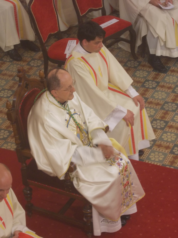 Eine Gruppe von Priestern in weißen Roben sitzt auf Stühlen auf einem roten Teppich in einer Kirche, einige halten Papiere.