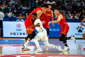 Eine Gruppe von Männern, die Basketball spielen, in einem Stadion, einer hält den Ball, mit Zuschauern im Hintergrund.