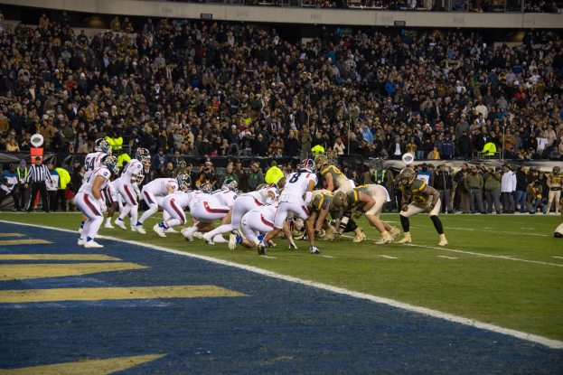 Ein Fussballspiel im Gange mit Spielern in Sportbekleidung und Helmen auf dem Feld, Zuschauern, Geländern und Texttafeln im Hintergrund.