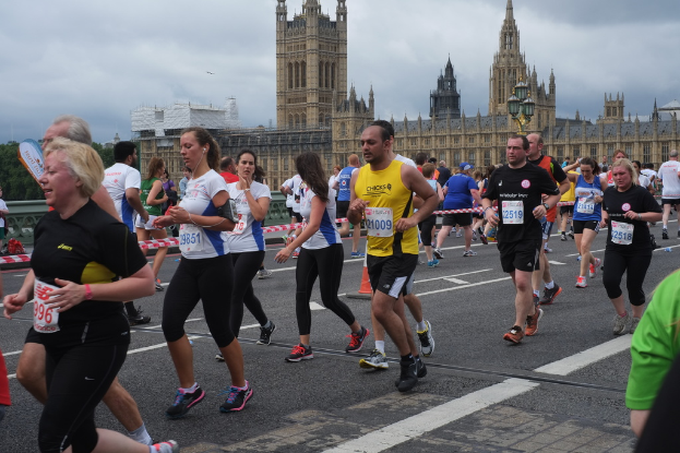 Eine Gruppe von Menschen, die mit einer Person in einem weißen und schwarzen Kleid an der Spitze laufen, vor einem cremefarbenen Gebäude, Bäumen und einem weißen Himmel.