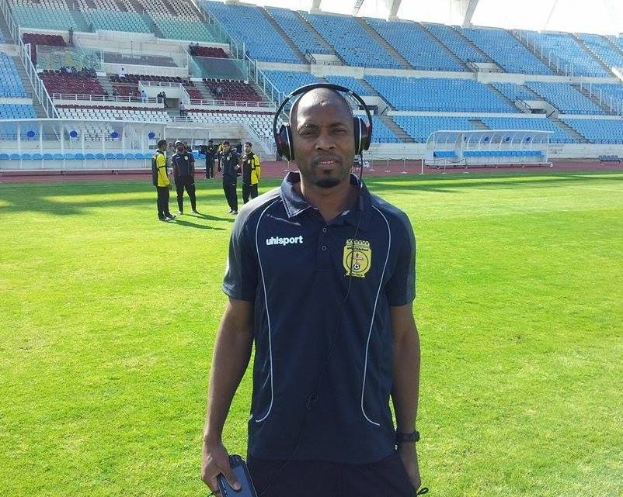 Ein Mann mit Headset, wahrscheinlich der Trainer der ghanaischen Nationalmannschaft, steht auf einem Fussballfeld in der Nähe eines Stadions und hält ein Objekt in der Hand, während im Hintergrund Menschen zu sehen sind.
