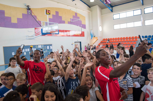 Eine Gruppe von Kindern steht vor einem Basketballfeld, einige halten Mobiltelefone, mit einer Tafel, Uhr, Torpfosten, Basketballkorb, Deckenbeleuchtung, Stühlen und Fenstern im Hintergrund.