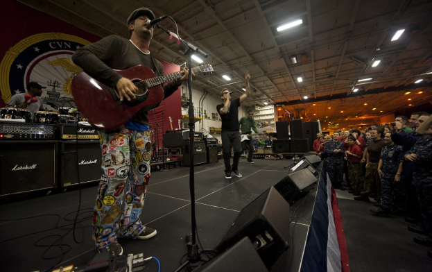 Ein als Chris Stapleton identifizierter Mann spielt eine E-Gitarre auf der Bühne, trägt eine Mütze, hält ein Mikrofon, sind Lautsprecher, eine Flagge und andere Instrumente in der Nähe, vor einem Publikum unter Deckenlampen mit einem Banner an der Wand dahinter.