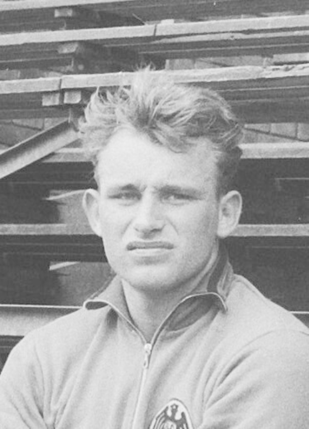 Schwarzes und weißes Foto eines Mannes in einer Jacke vor einem Haufen Holzplanken, der als legendärer deutscher Fußballspieler gilt.