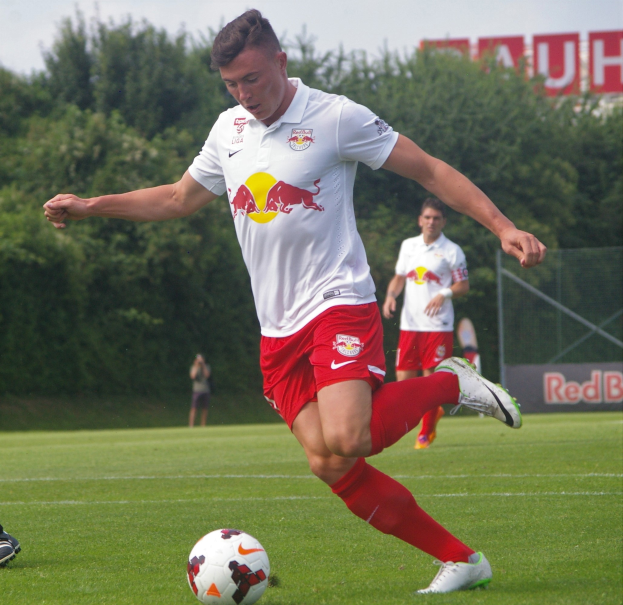Ein Mann in einem Fußballtrikot schießt einen Ball auf einem grünen Feld mit Bäumen und einem klaren blauen Himmel im Hintergrund, wobei ein Schild mit der Aufschrift 'RB Leipzig' sichtbar ist.