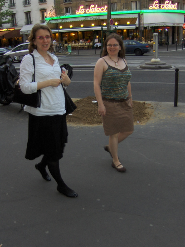 Zwei Frauen auf einer Straße mit vielen Fahrzeugen, Gebäuden mit Text, Lichtern, einem Baum und kleinen Maulwürfen.