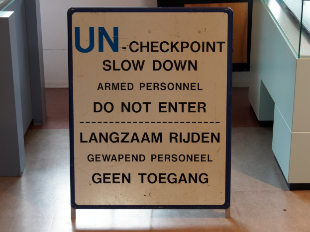 Schild auf dem Boden mit der Aufschrift "UN-Checkpoint Verlangsame bewaffnete Personen - Betreten verboten" mit einem Glastisch und Gegenständen rechts und einer Wand mit einem Gegenstand im Hintergrund.