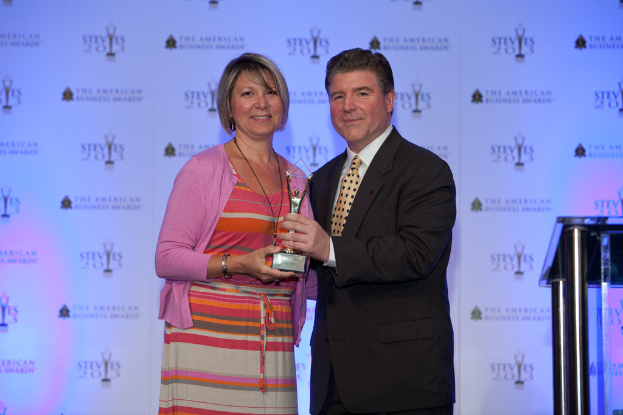 Zwei Personen mit einer Trophäe, wobei eine Person einen schwarzen Blazer und ein weißes Hemd trägt und ein Banner im Hintergrund zu sehen ist.