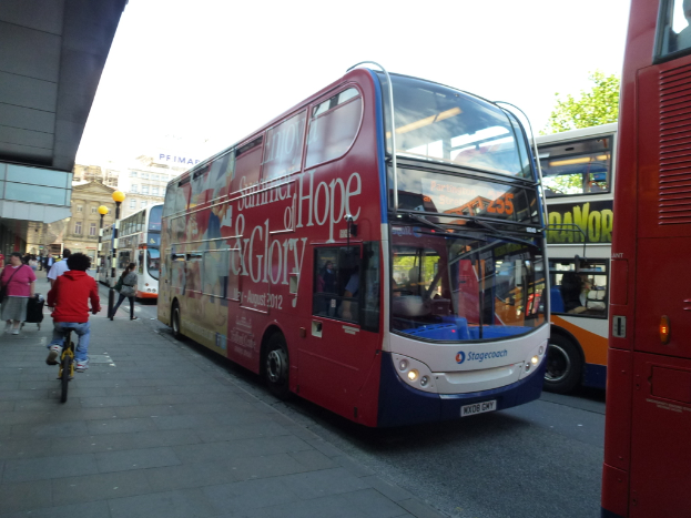 Eine Person fährt mit dem Fahrrad auf einem Gehweg mit vielen Menschen, Bussen mit Text darauf, Gebäuden und Laternen im Hintergrund.