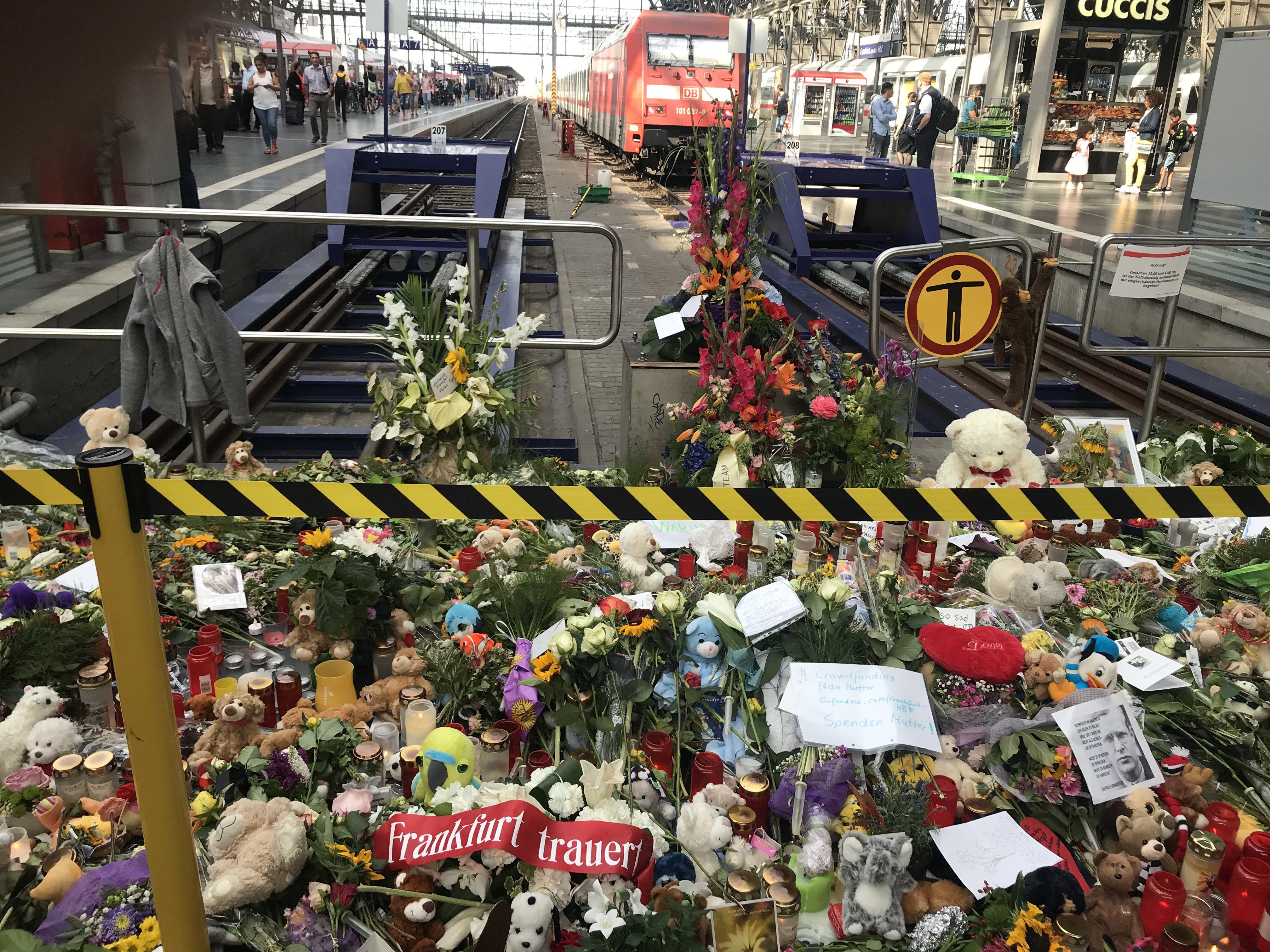 Ein Bahnsteig mit einem Zug im Hintergrund, umgeben von Menschen und verschiedenen Gegenständen wie Teddybären, Blumensträußen und Kerzen, die an ein Unglück in Berlin erinnern.