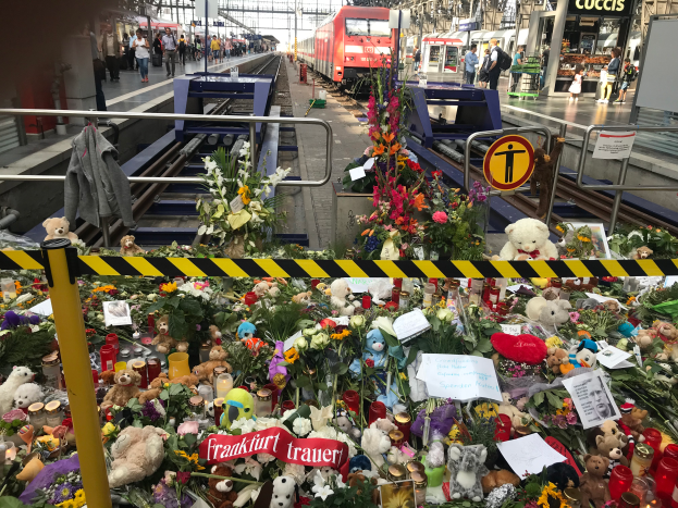 Ein Bahnsteig mit einem Zug im Hintergrund, umgeben von Menschen und verschiedenen Gegenständen wie Teddybären, Blumensträußen und Kerzen, die an ein Unglück in Berlin erinnern.