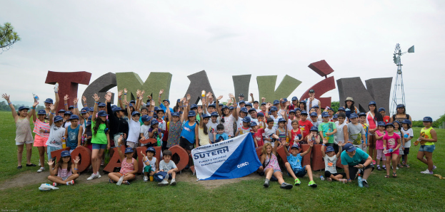 Mehrere Kinder stehen draußen auf Gras, halten die Hände hoch und posieren für ein Bild, einige halten ein Banner, mit Blöcken dahinter, einem Pfahl rechts, Bäumen links und dem Himmel darüber.