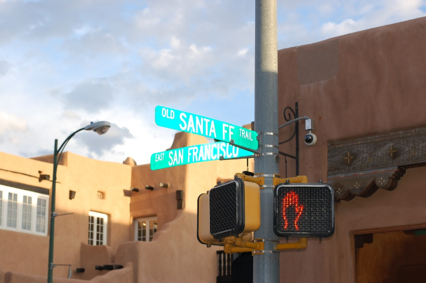 Ein Mast mit angebrachten Namensschildern und -schildern, Straßenlaternen, Gebäuden mit Fenstern und einem bewölkten Himmel im Hintergrund.