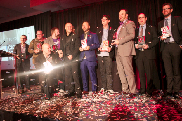 Eine Gruppe von Jungs steht auf einer Bühne und hält Schilde, mit einem Mann in der Mitte, der das Licht reflektiert, einem Podium mit einem Mikrofon auf der linken Seite, einem Bildschirm und einem schwarzen Vorhang im Hintergrund sowie verstreuten Papierstücken auf der Bühne.