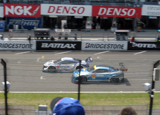 Zwei Sportwagen rasen auf einer Rennstrecke mit einer Zuschauermenge hinter einem Maschendrahtzaun, Pfosten und Lichtern, mit Texttafeln im Hintergrund.