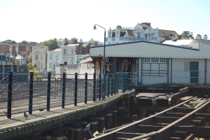 Ein Bahnhof mit einem Zug auf den Schienen, eine Brücke mit Geländern, Laternenmasten, Verkehrsampeln mit Masten, Gebäude mit Fenstern, Bäume und ein Himmel im Hintergrund.