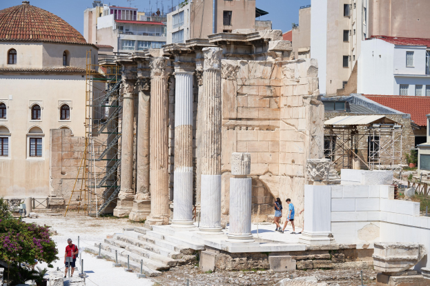 Ruinen des Tempels von Olympian Zeus in Athen, Griechenland, mit Besuchern zwischen verstreuten Steinen und Vegetation unter einem bewölkten Himmel.