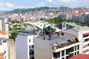 Eine Stadtansicht von einem hohen Standpunkt aus, die zahlreiche Gebäude, Bäume, Straßen, Fahrzeuge und entfernte Berge unter einem bewölkten Himmel zeigt, mit Solarpanelen auf dem Dach eines Gebäudes.