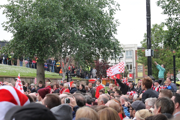 Eine Menge Menschen, einige halten Fahnen, steht auf einer grasbewachsenen Fläche vor einem mehrstöckigen Gebäude mit Fenstern, mit einem Lichtmast und Bäumen im Vordergrund.